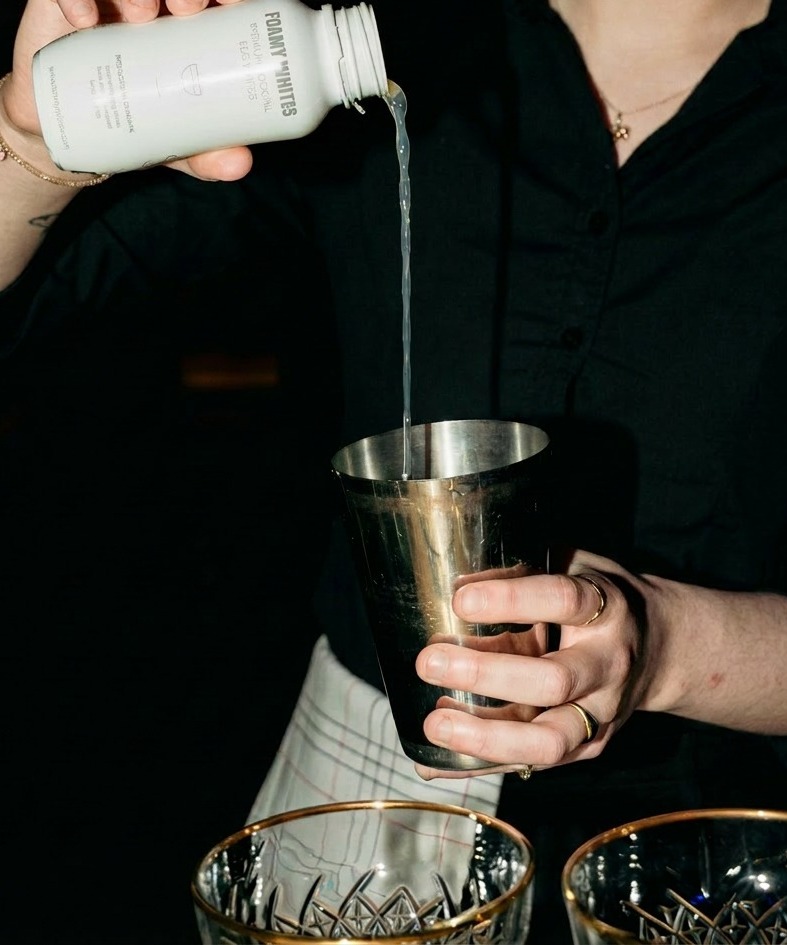 A bartender pours a single measure of FOAMY WHITES from the bottle into a silver cocktail shaker