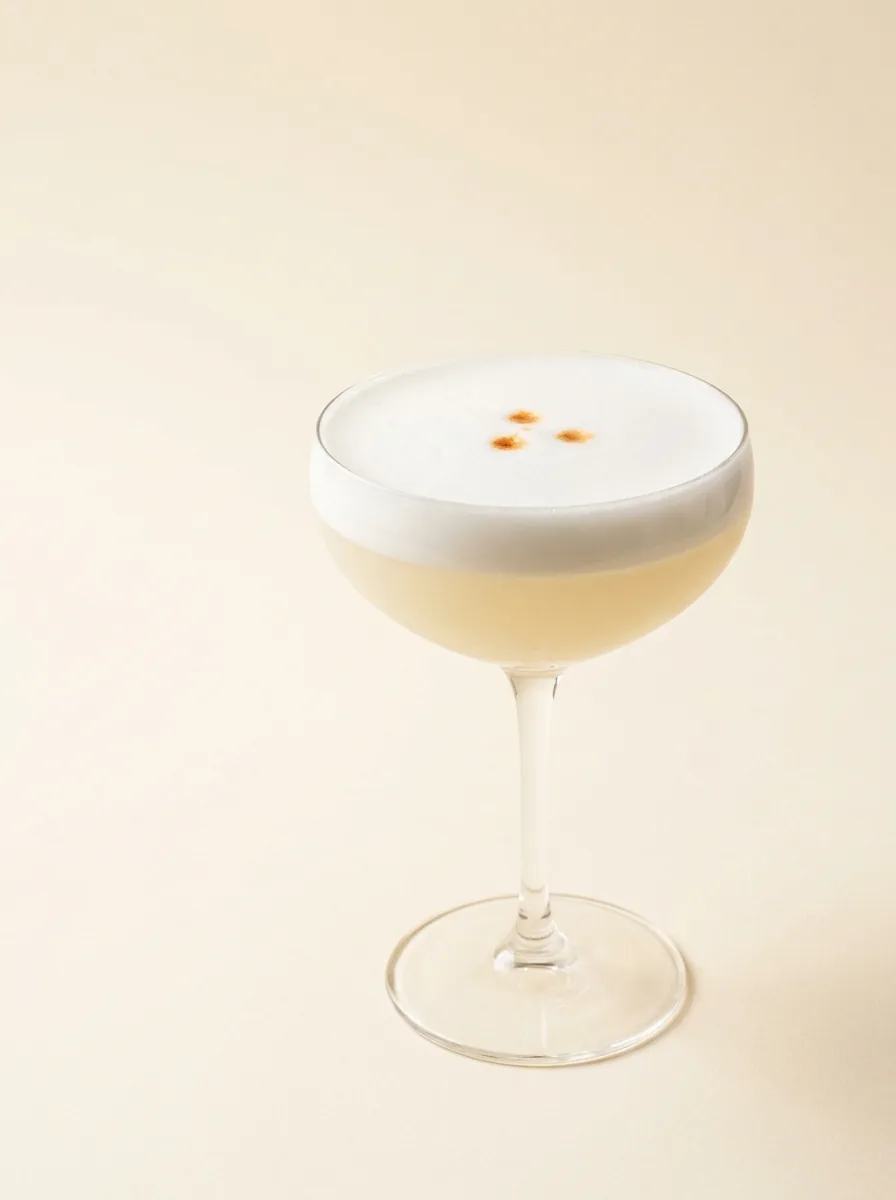 Pisco Sour served in a coupe with a dense white foam finished with three drops of Angostura bitters, photographed on a warm off-white surface.